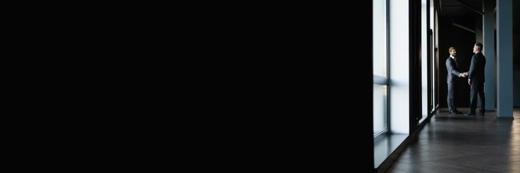 Two executives in business suits shake hands in a glass‑walled office hallway—symbolizing Scorpion Five Technologies’ partnership with large enterprises and government clients to tackle complex technology challenges in enterprise systems, cloud engineering, hybrid cloud, and cybersecurity accreditation.