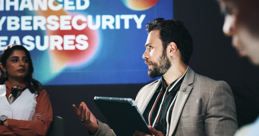 Business professionals discussing cybersecurity compliance strategies during a meeting, with a presentation screen in the background displaying the words "Enhanced Cybersecurity Measures."