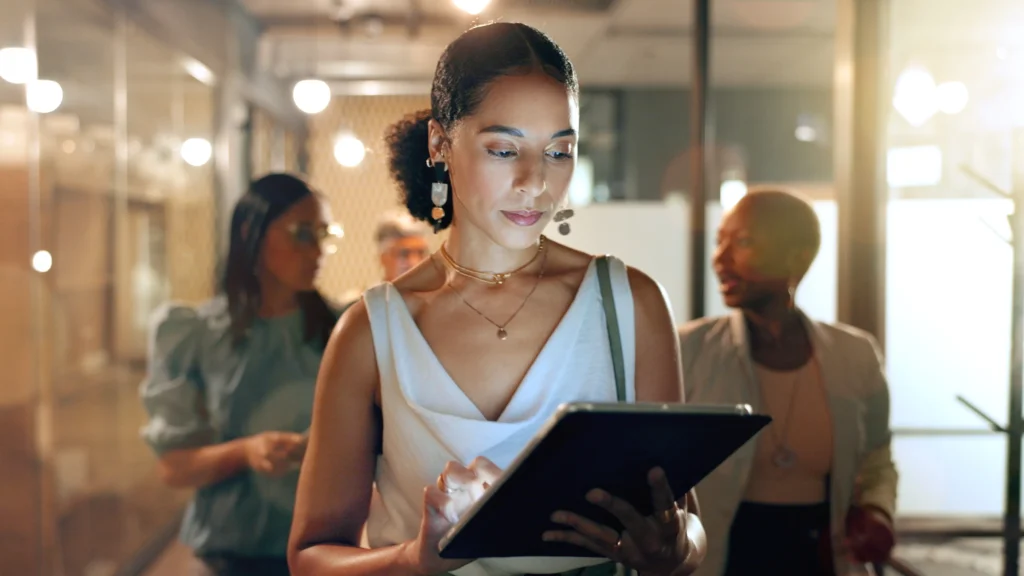 Focused professional using a tablet while colleagues talk in the background, symbolizing the importance of avoiding distractions and staying on track with digital marketing priorities.