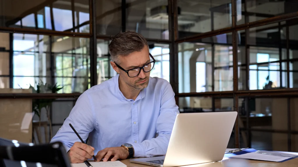 Business professional analyzing notes while working on a laptop, representing the process of assessing current digital marketing efforts and identifying gaps.