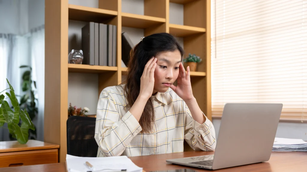 Stressed small business owner sitting at a desk with laptop, holding her head in frustration while struggling with online marketing tasks.