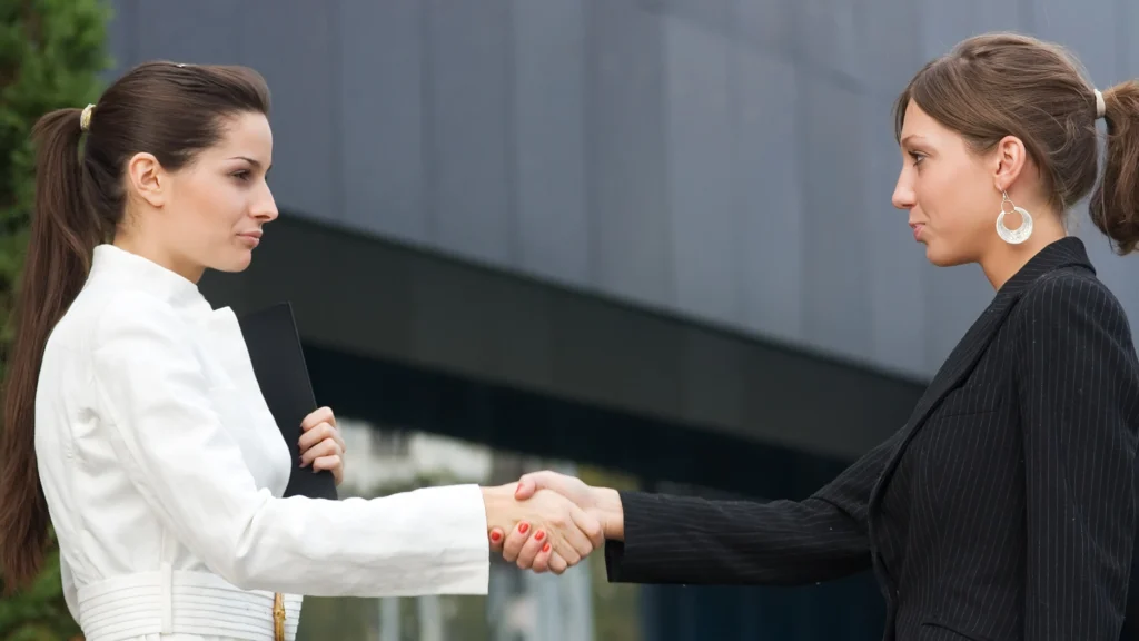 Two business professionals shaking hands while glaring suspiciously at one another outside an office building, representing competition, rivalry, and the importance of understanding competitors in digital marketing.